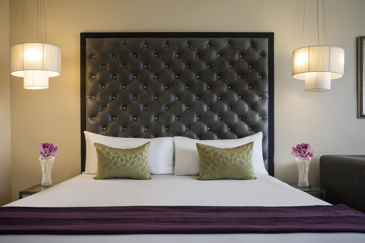 Una habitación de hotel con cabecera capitonada, dos lámparas de noche, una cama con ropa blanca y dos jarrones decorativos con flores.
