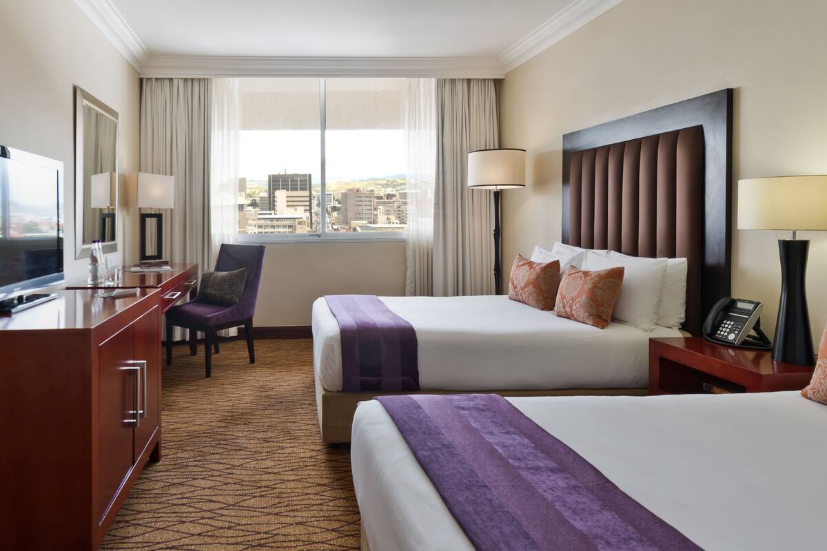 A hotel room with two beds, a desk with a TV, a chair, and a view of the city.