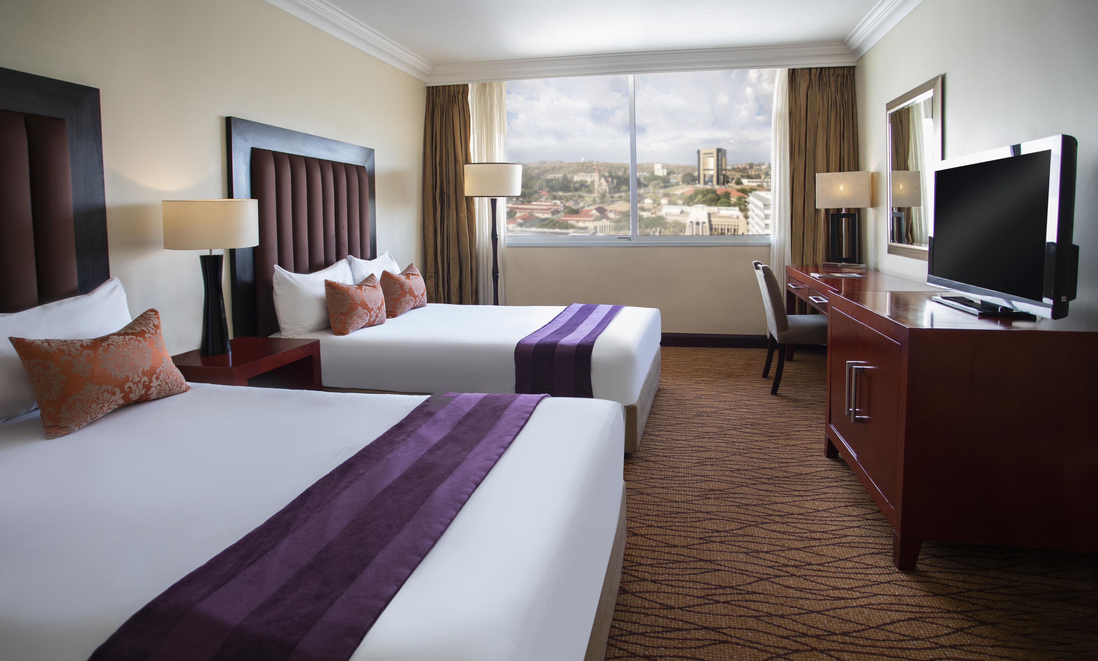 A hotel room with two beds, a television, a desk, and a view of the city.