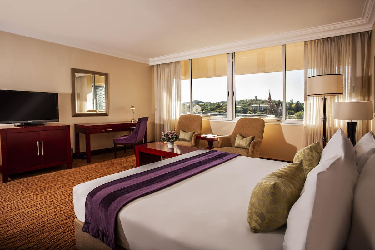 A hotel room with a large bed, a desk, a television, and a view of a building through the window.
