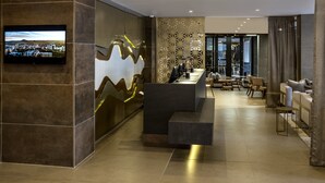 Un moderno vestíbulo de hotel con una recepción, una zona de estar y una gran pantalla de televisión que muestra un paisaje urbano.