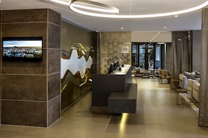 Un moderno vestíbulo de hotel con una recepción, una zona de estar y una gran pantalla de televisión que muestra un paisaje urbano.