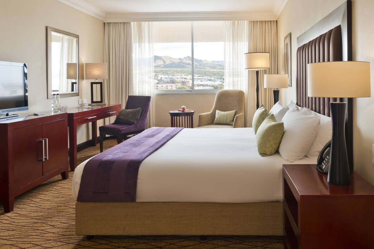 A hotel room with a large bed, a desk with a TV, a chair, a small table, and a view of mountains through the window.