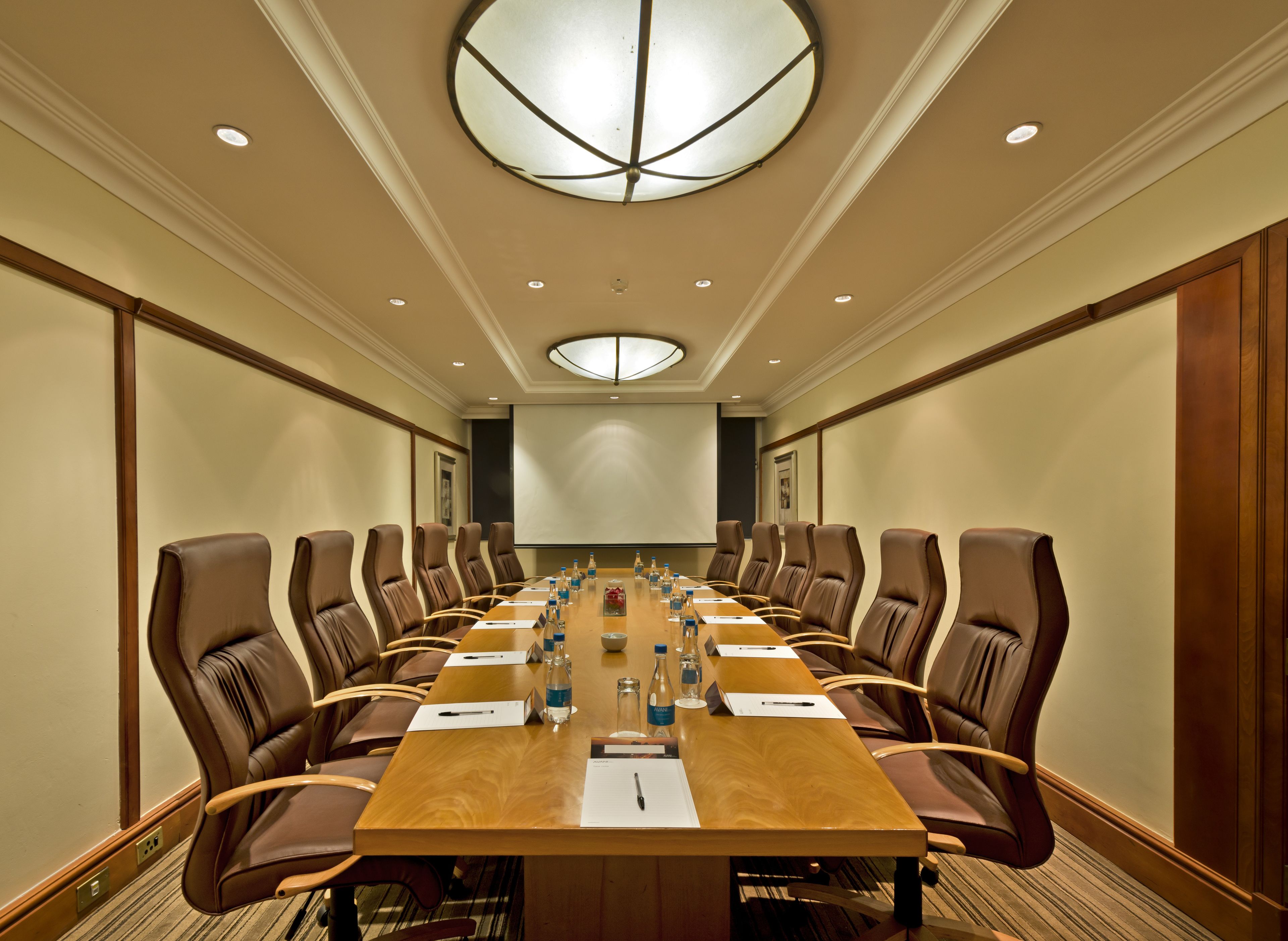 A conference room with a long table, brown leather chairs, and a large screen at the front.
