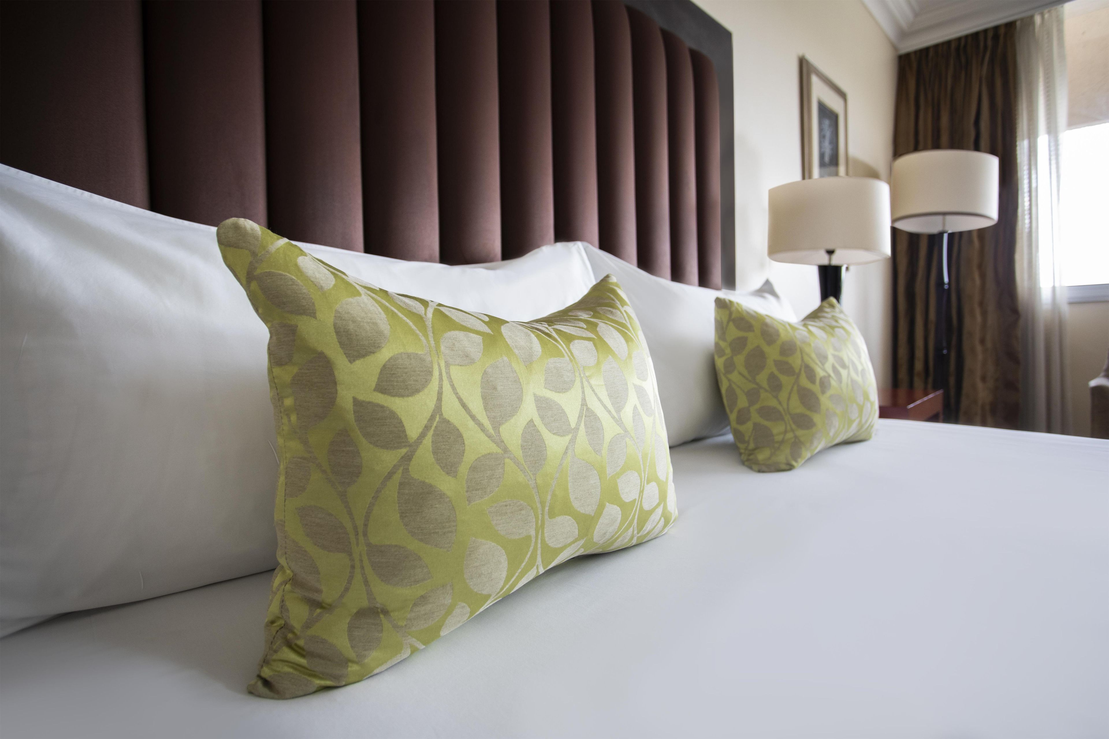 Una habitación de hotel con una cama grande, dos almohadas con estampado de hojas, un cabecero, una lámpara y un cuadro en la pared.