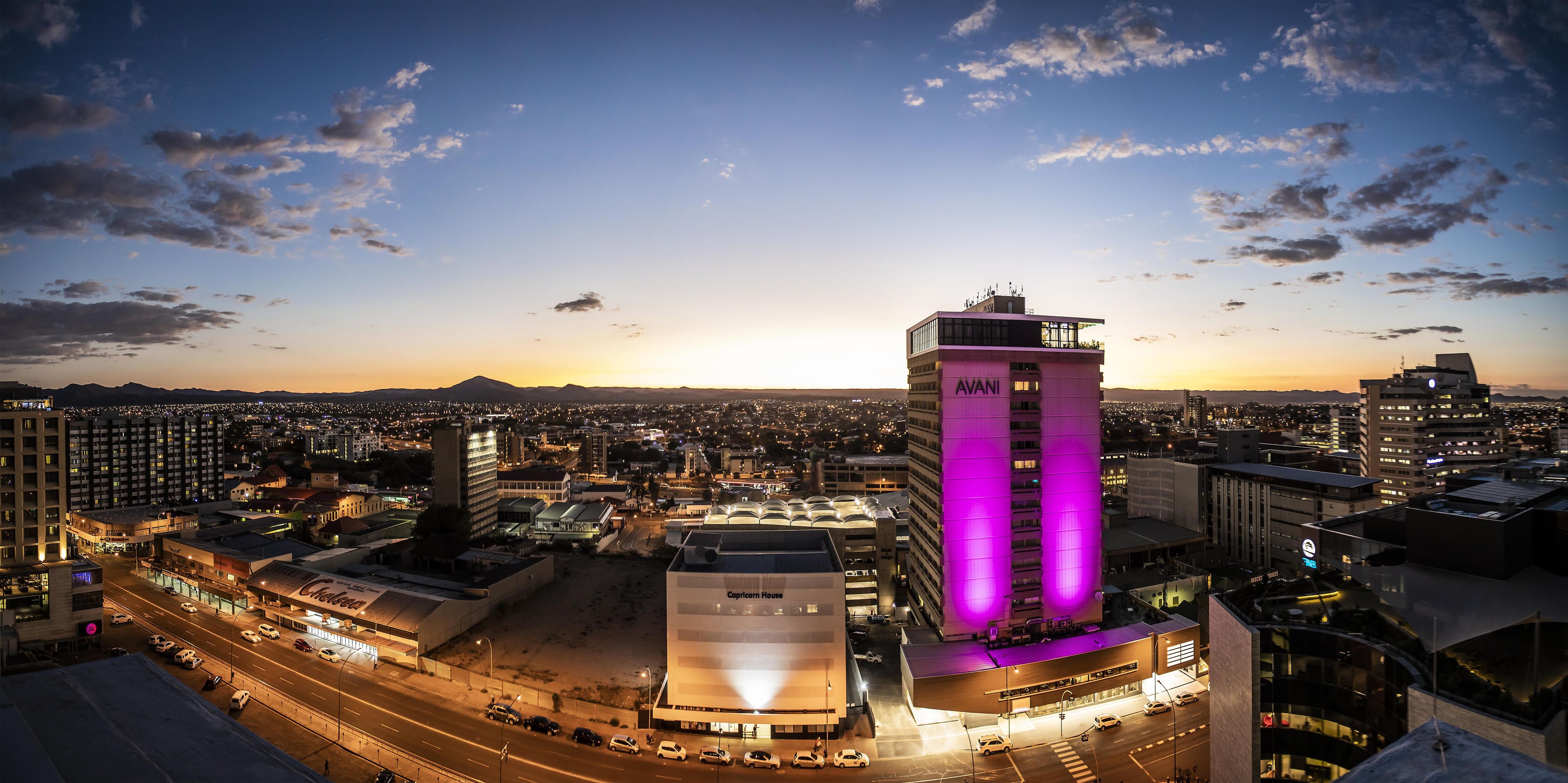 A cityscape at dusk with prominent buildings including a tall structure with purple lighting and a building with a sign that reads 'Capricorn House'.