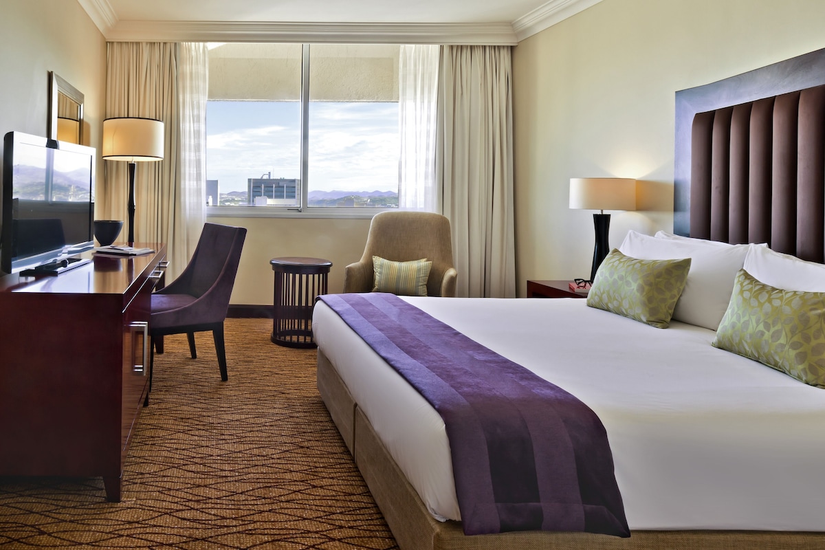 A hotel room with a large bed, a desk with a TV, a chair, and a view of the city through the window.