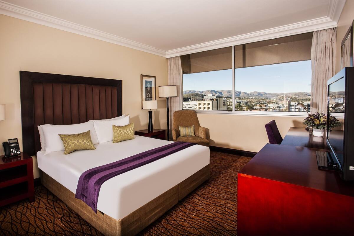 A hotel room with a large bed, a desk, a chair, a lamp, a phone, and a view of the city.
