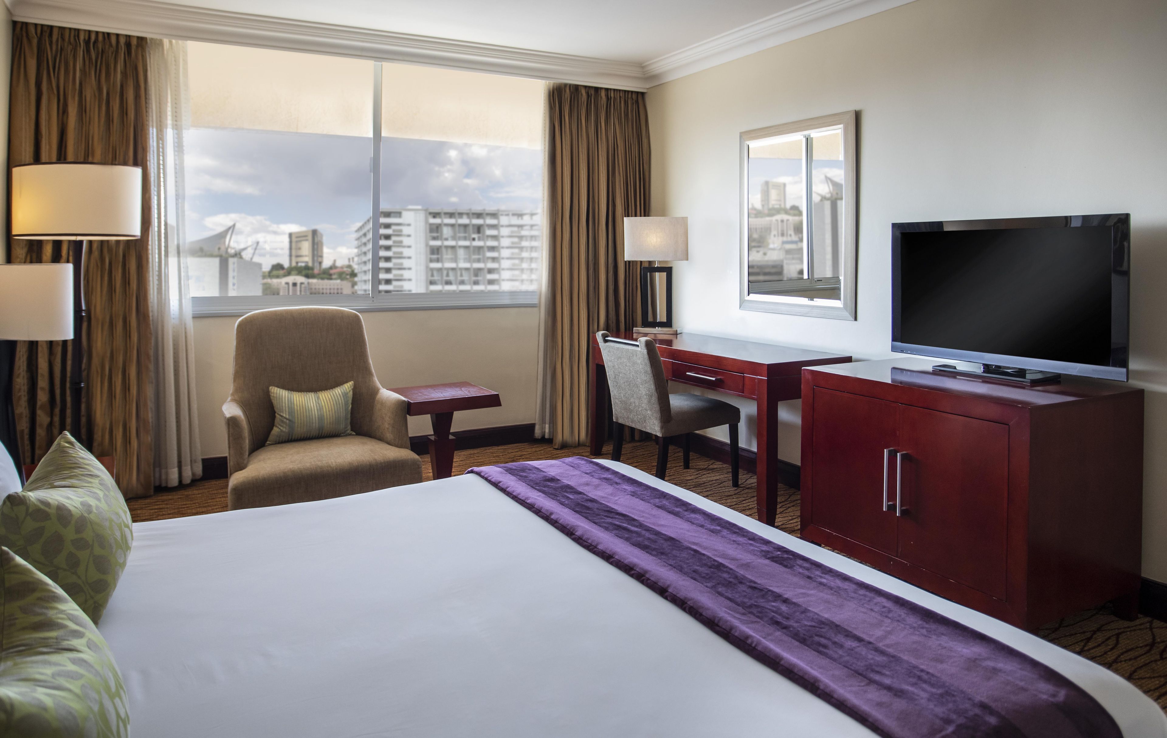 A hotel room with a bed, a desk, a chair, a television, and a view of the cityscape through the window.