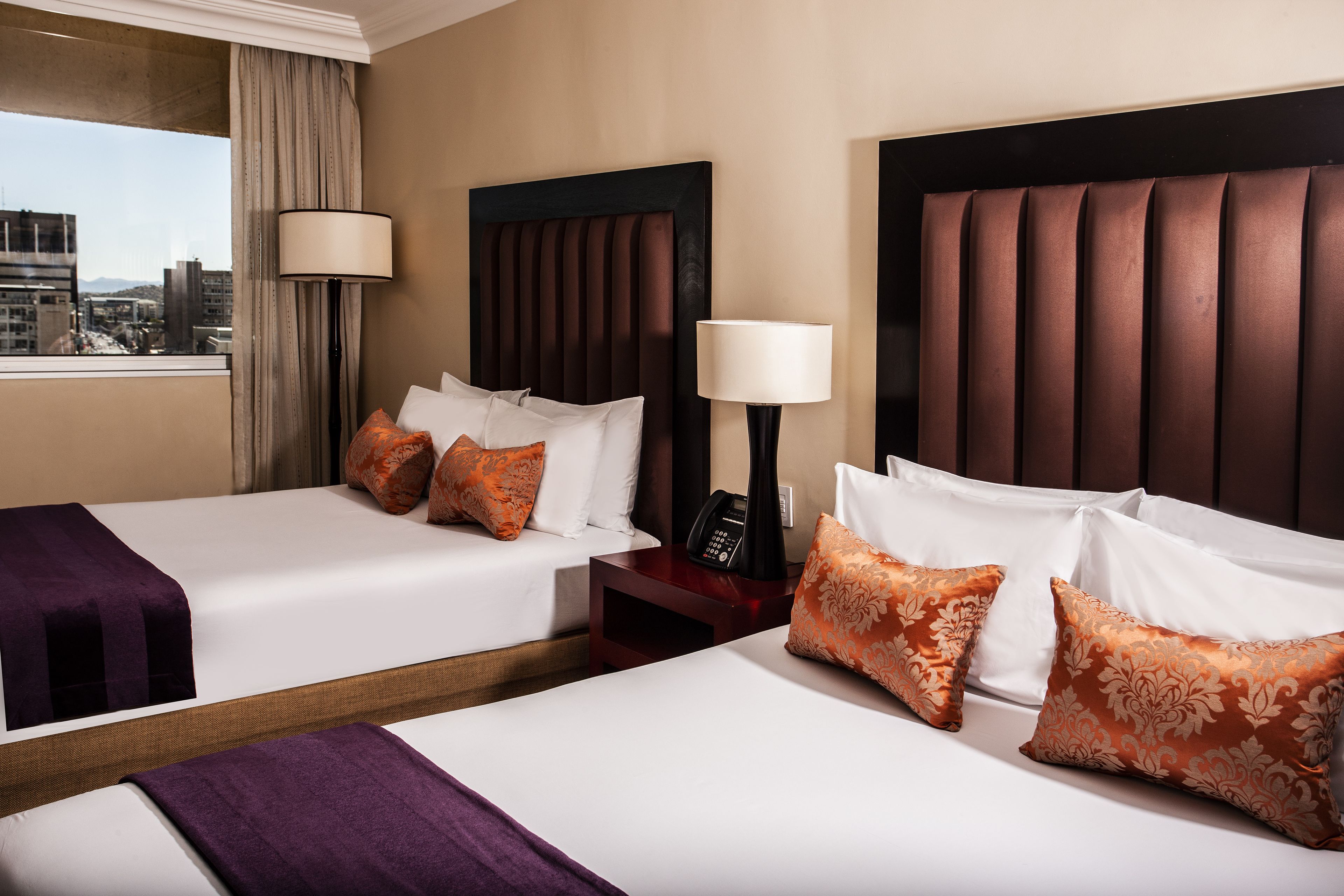 A hotel room with two beds, each with white linens and decorative pillows, a nightstand with a phone, and a window with a view of buildings.