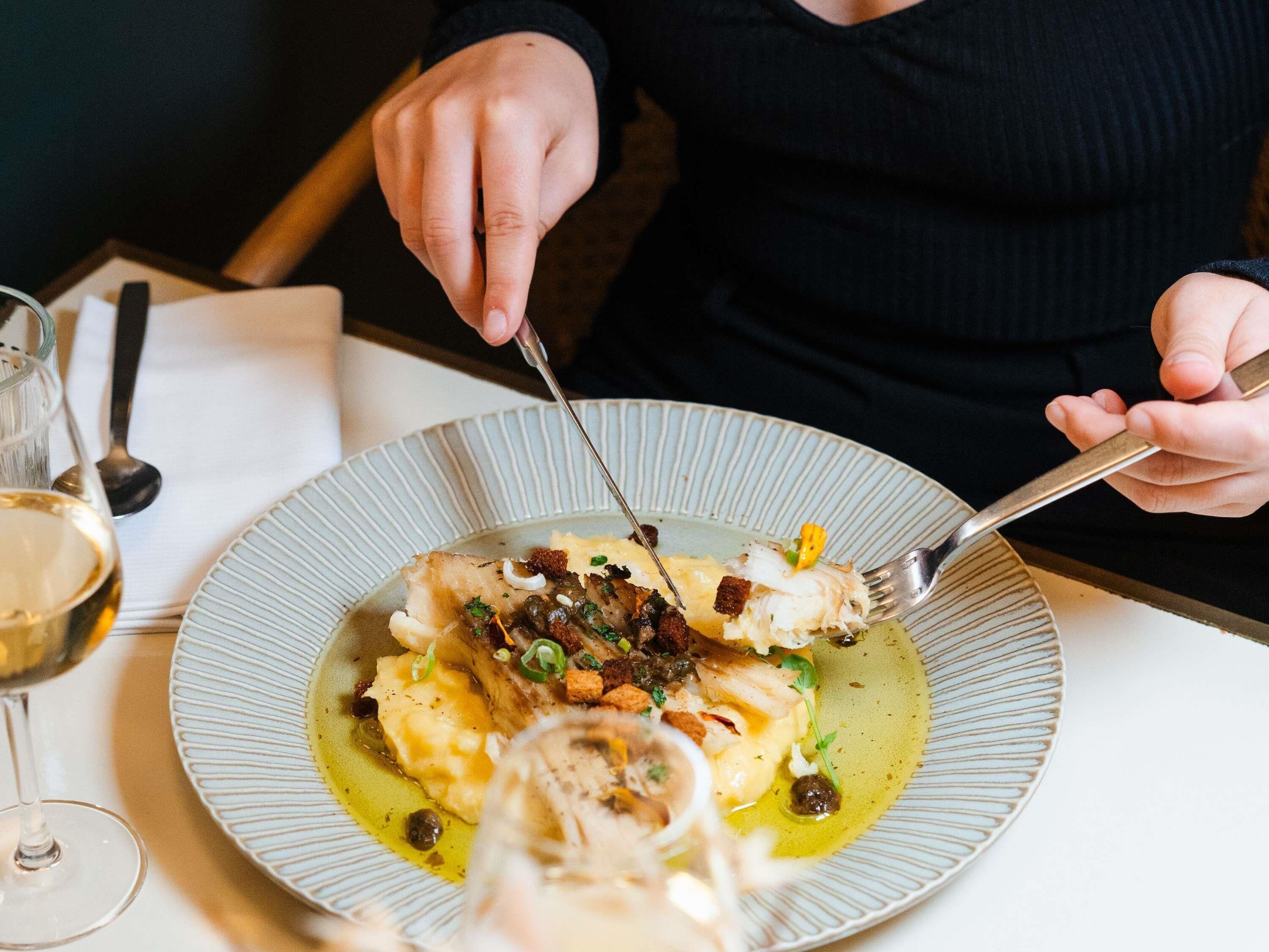 A person dining at a table with a plate of food, a glass of wine, and a fork.