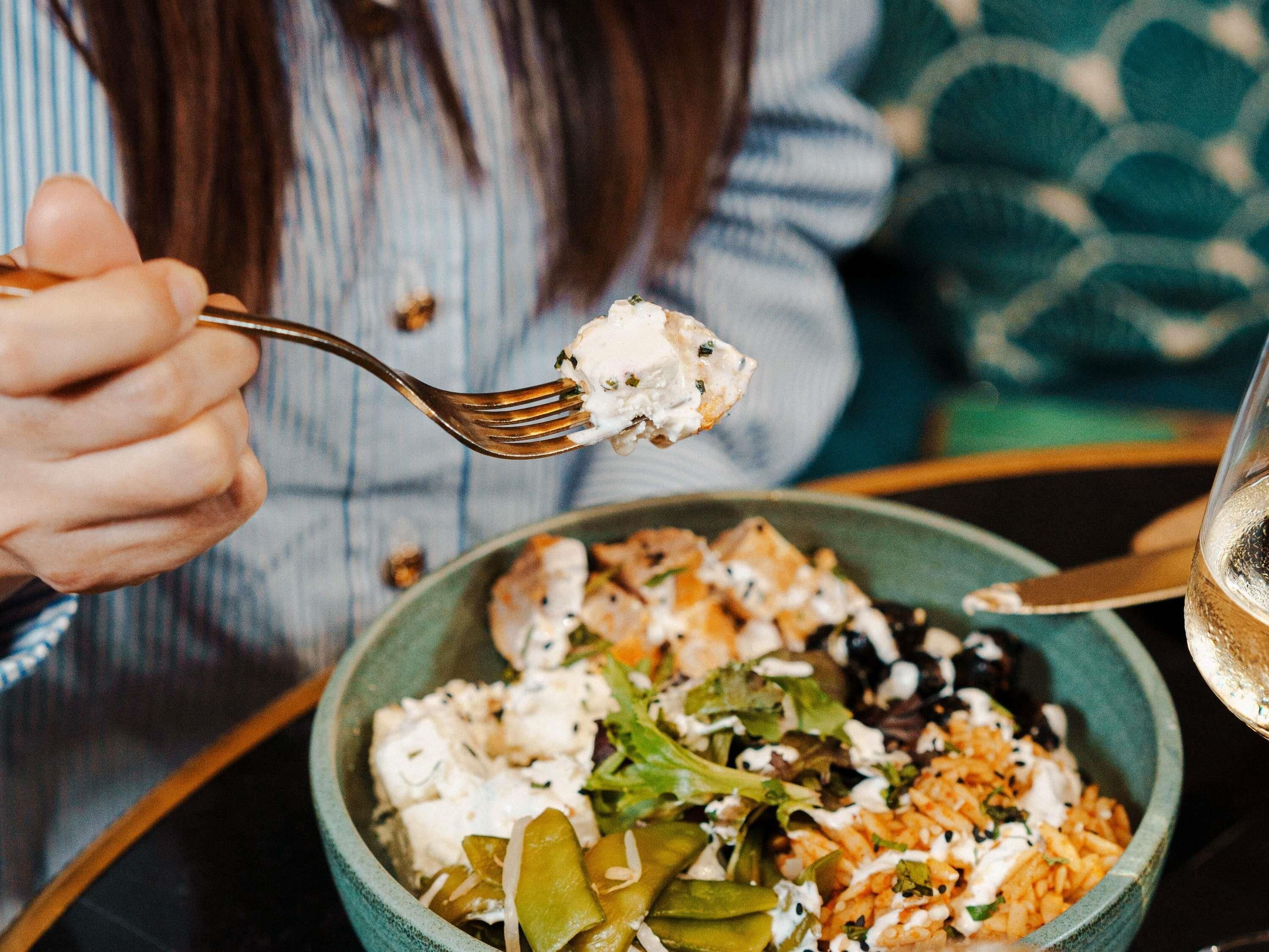 A person holding a fork with a creamy substance, a bowl of food with various ingredients, and a glass of a clear liquid on a table.