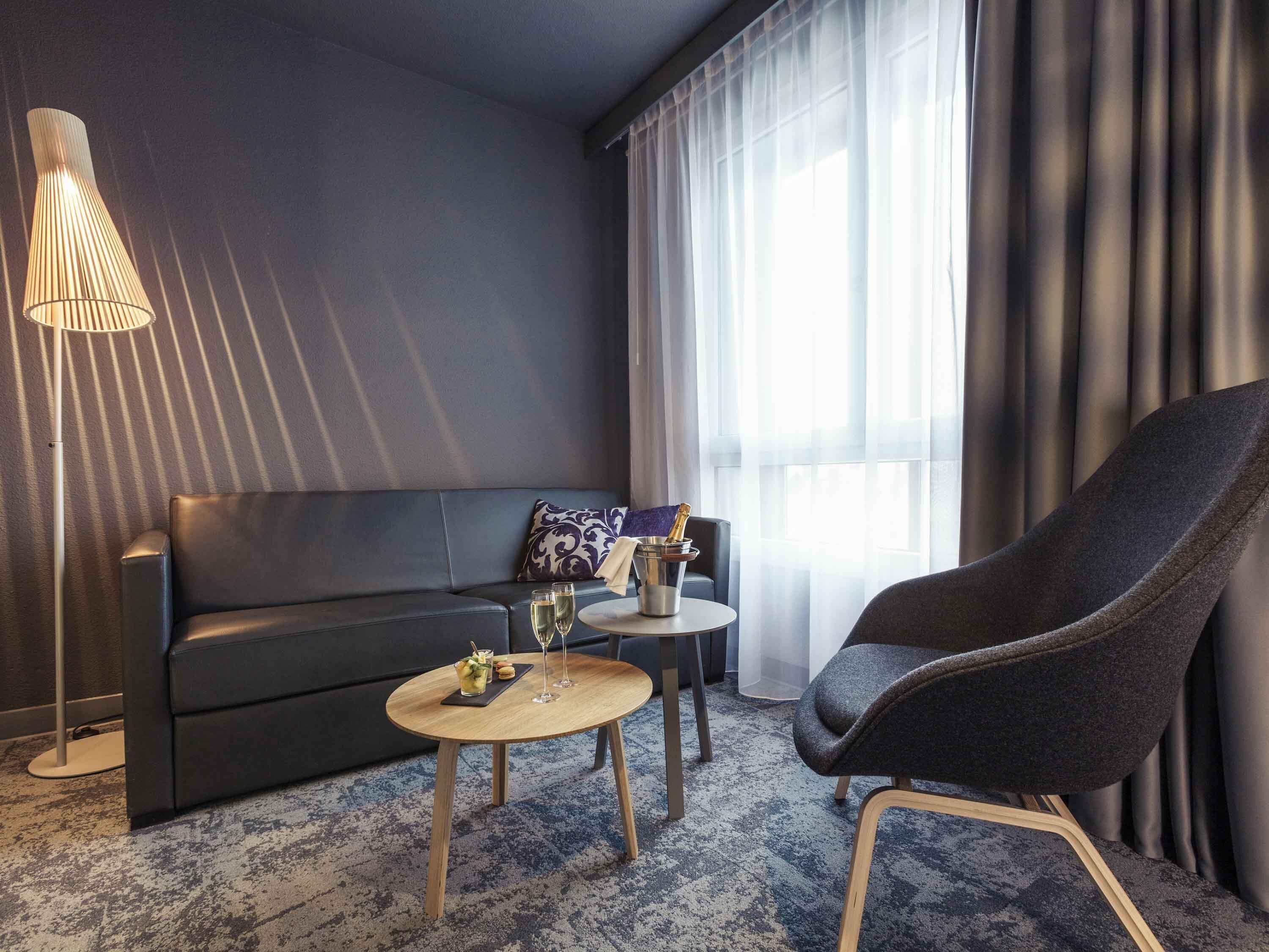 Quarto de hotel moderno com um sofá de cor escura, uma mesa de centro redonda de madeira, uma poltrona escura, um abajur e uma janela com cortinas translúcidas.