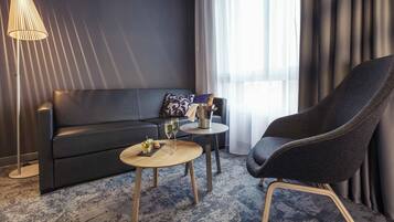 A modern hotel room with a dark-colored sofa, a round wooden coffee table, a dark armchair, a floor lamp, and a window with sheer curtains.