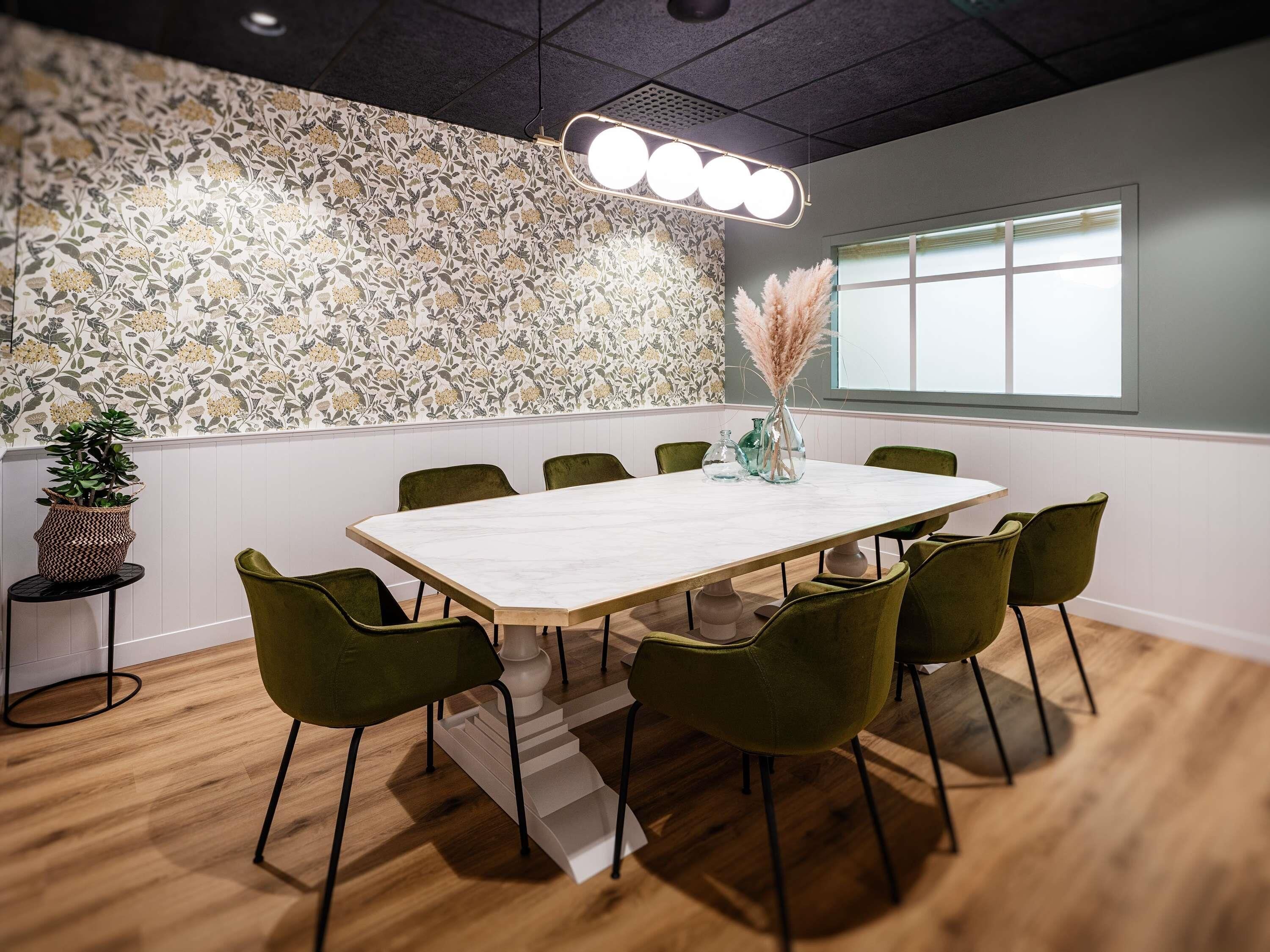 A conference room with a floral wallpaper, a white table, green chairs, and a vase with pampas grass.