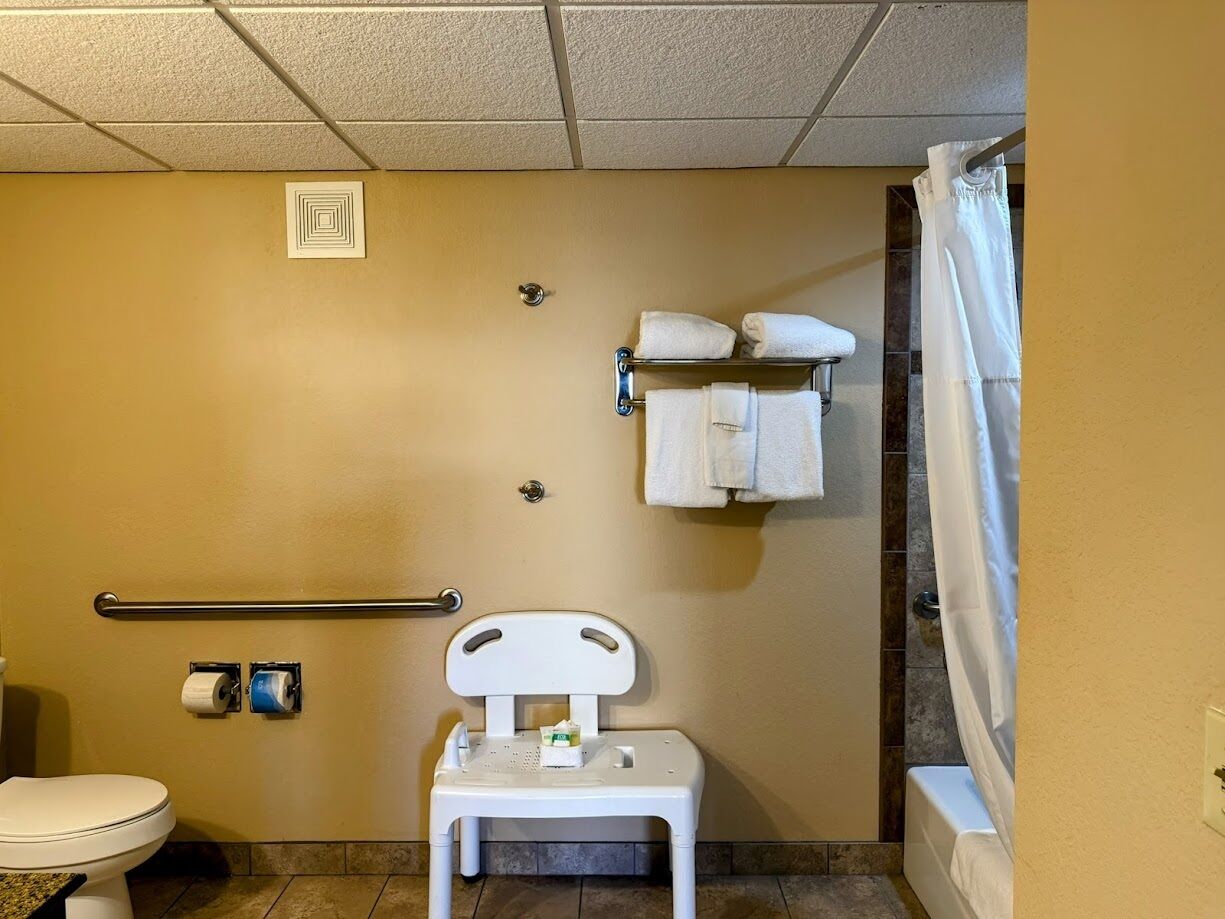 Un baño pequeño y funcional con inodoro, asiento de inodoro elevado, barra de apoyo, toallero con toallas y cortina de ducha.