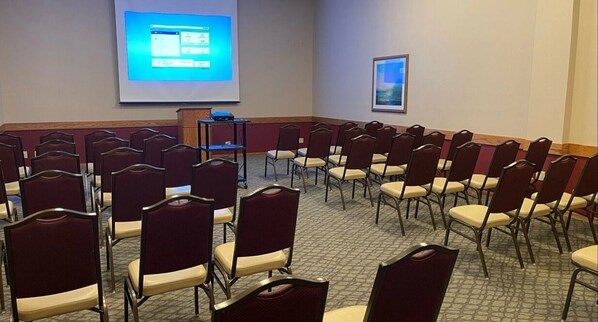 Un salón de conferencias con filas de sillas, una pantalla para proyector y una zona para presentaciones.