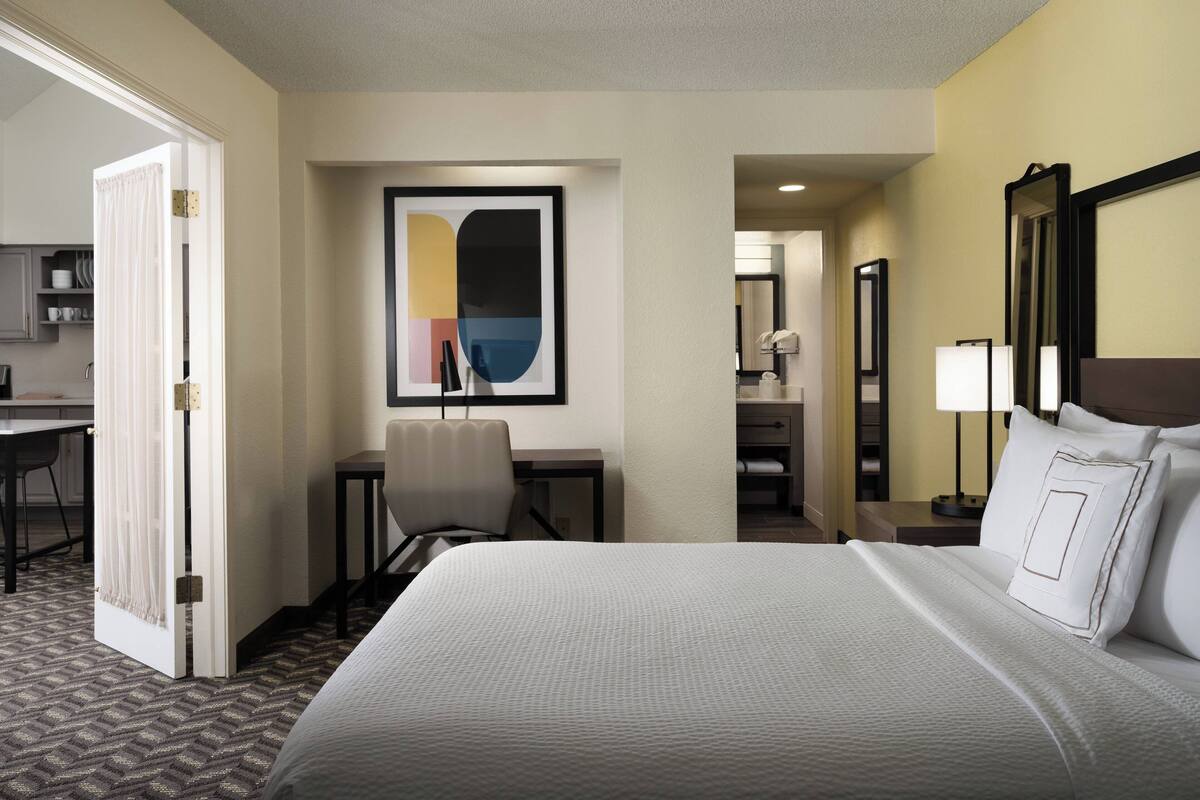 Une chambre d’hôtel avec un grand lit, un bureau, une chaise et une salle de bain visible à travers une porte ouverte.