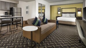 A hotel room with a kitchenette, a sofa with decorative pillows, a bed with a headboard, and a small table.