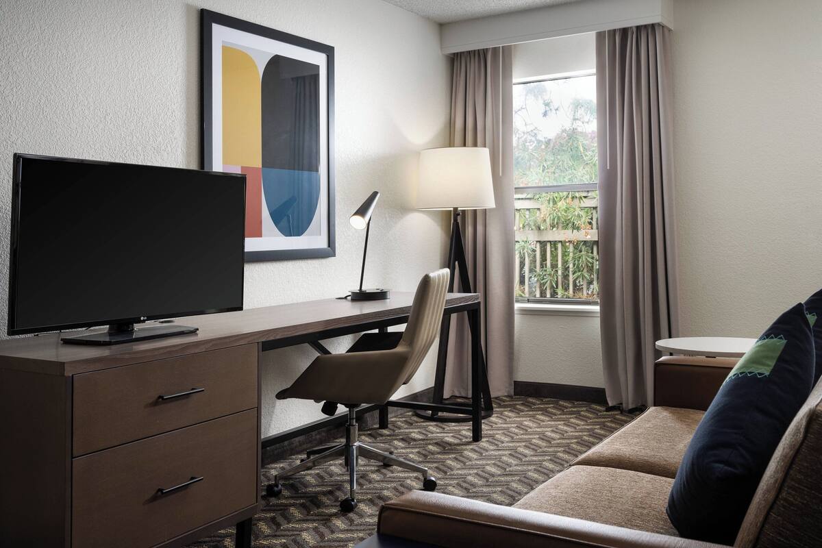 Une chambre d’hôtel équipée d’un bureau, d’un téléviseur à écran plat, d’une lampe, d’une chaise, d’un canapé et d’une fenêtre avec des rideaux.