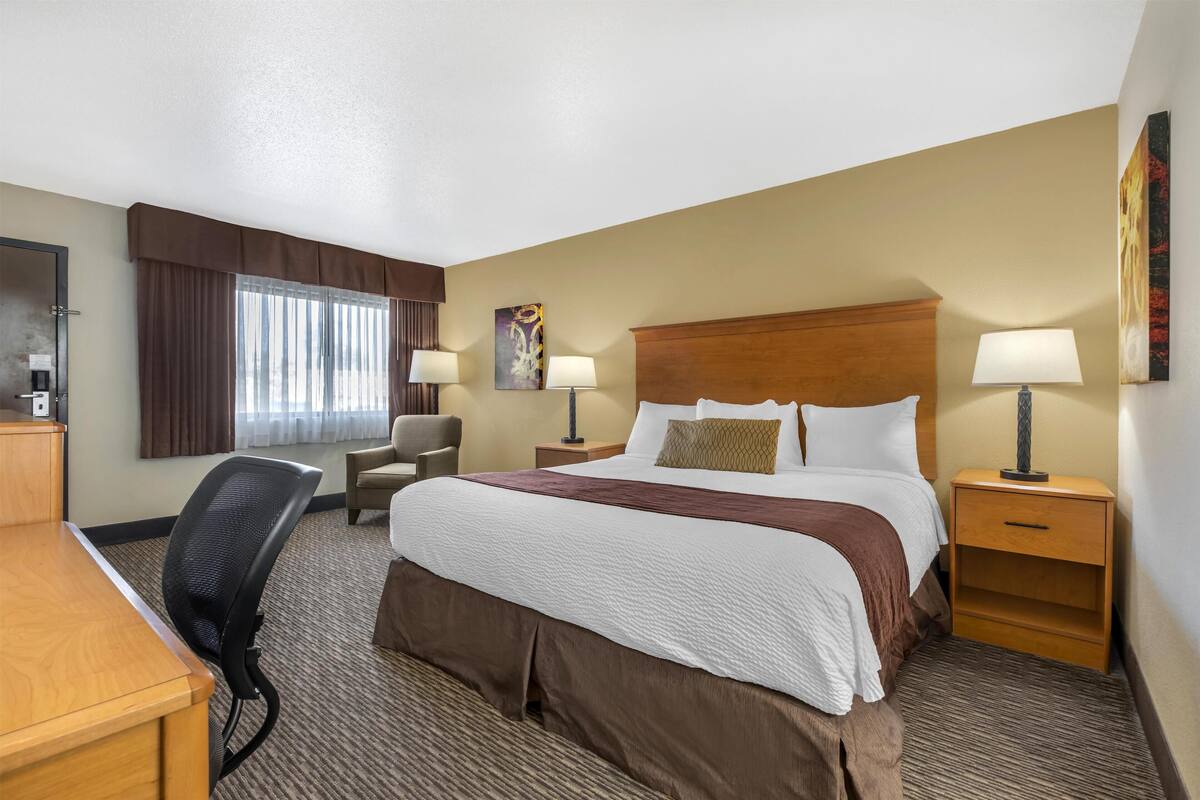 A hotel room with a large bed, two bedside tables with lamps, a desk, a chair, and a window with curtains.
