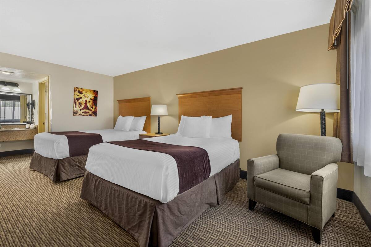 A hotel room with two beds, a chair, a desk, and a lamp.