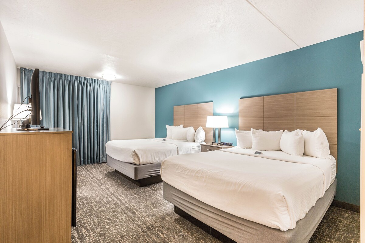 A hotel room with two beds, a desk, a lamp, and a view of the outdoors.