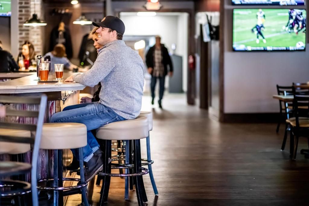 Una persona sentada en una barra con taburetes, una pantalla de televisión mostrando un evento deportivo, y una zona de bar con taburetes y mostrador.