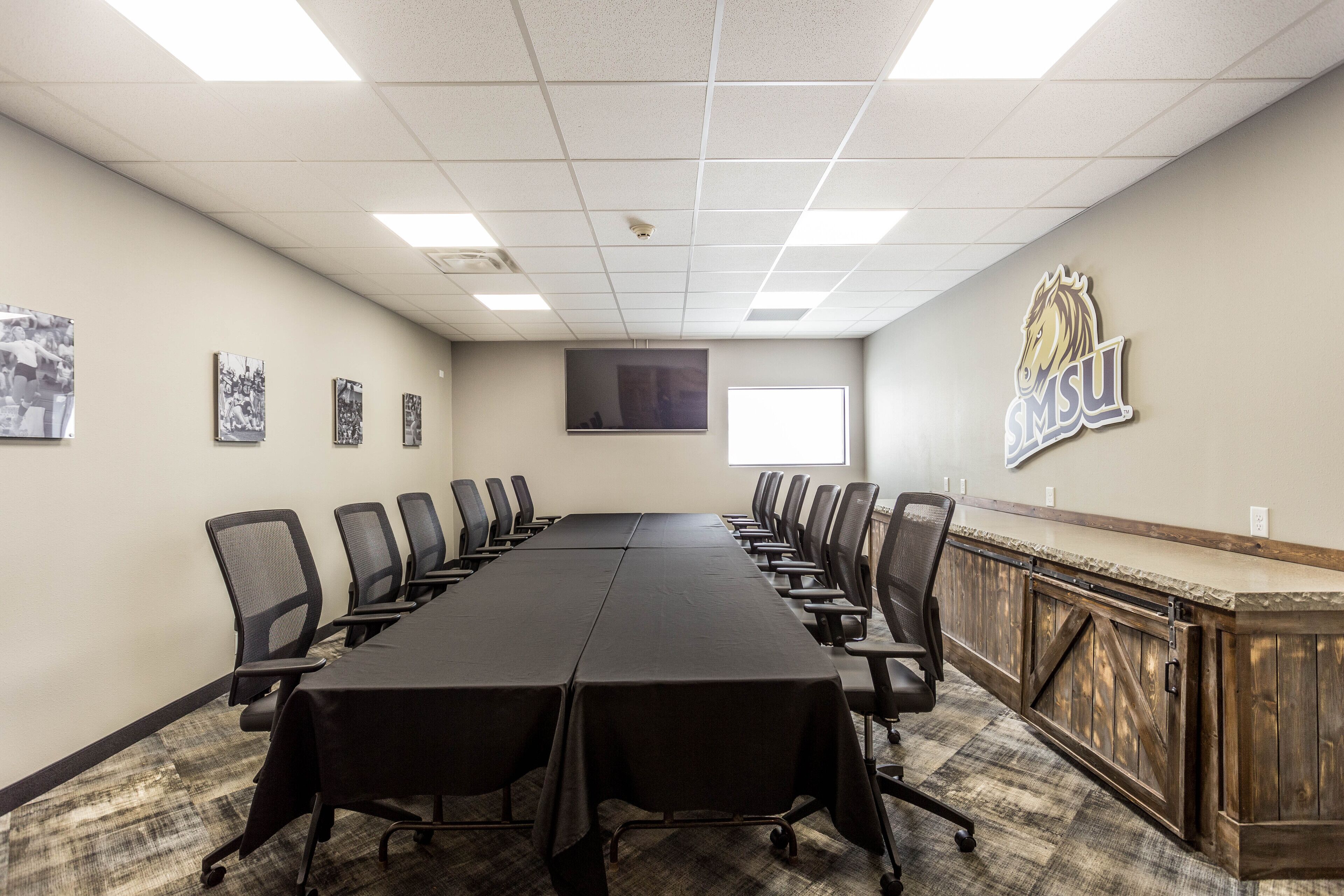 A conference room with a long table, chairs, a wooden cabinet, and a mounted television.