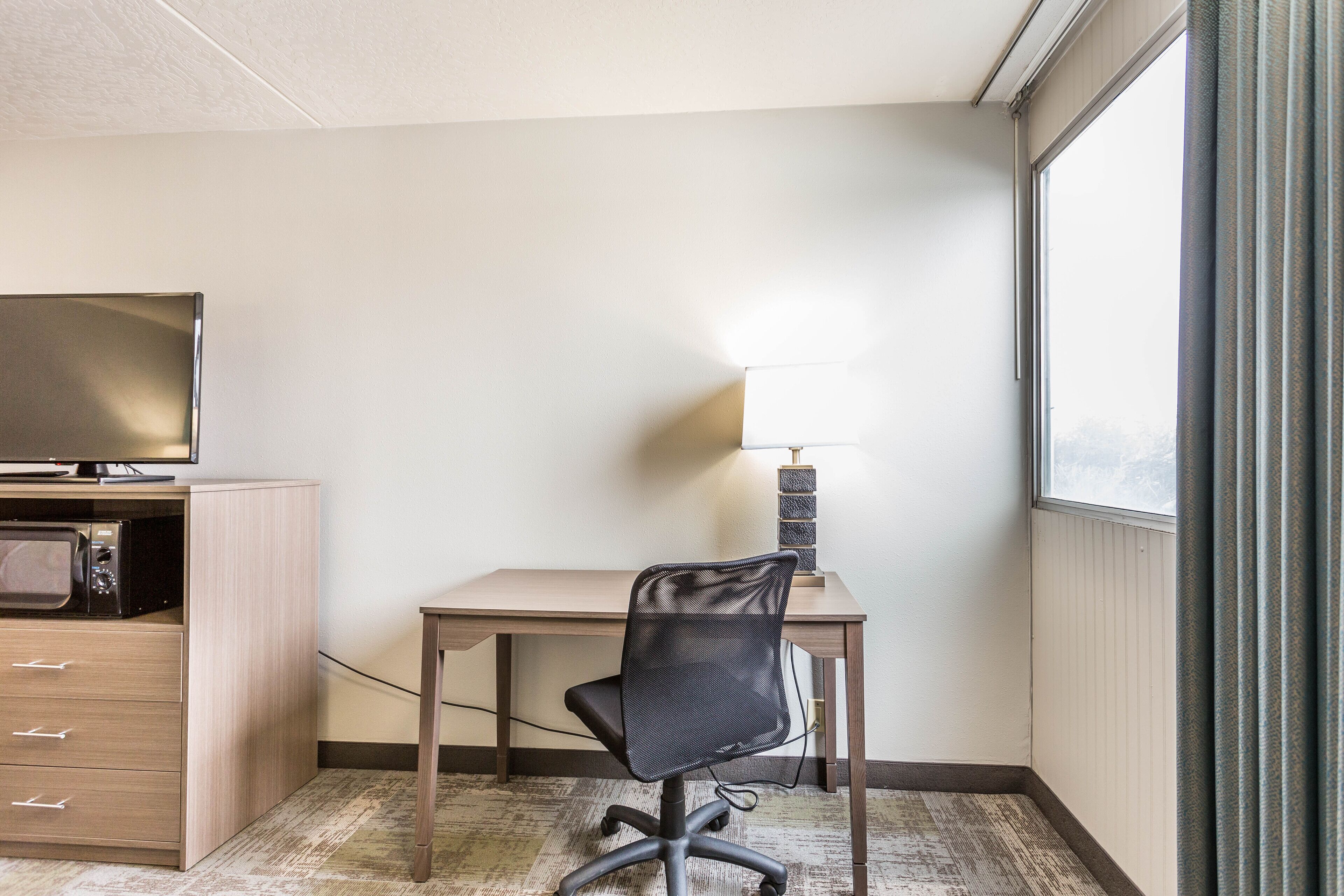 A hotel room with a desk, chair, TV, and a lamp.