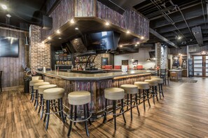 Uma área de bar com um balcão comprido, vários banquinhos e uma tela de televisão.