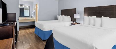 A hotel room with two beds, a desk, a chair, a TV, a bathroom with a mirror, and a nightstand with a lamp.