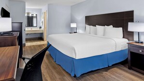 A hotel room with a large bed, a desk, a chair, a nightstand with a lamp, and a bathroom with a mirror and sink.