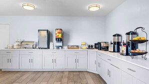 A modern kitchen with white cabinets, a coffee machine, a juice dispenser, and a fruit basket.