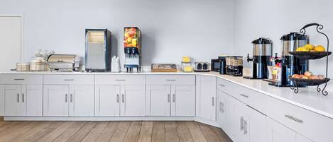 A modern kitchen with white cabinets, a coffee machine, a juice dispenser, and a fruit basket.