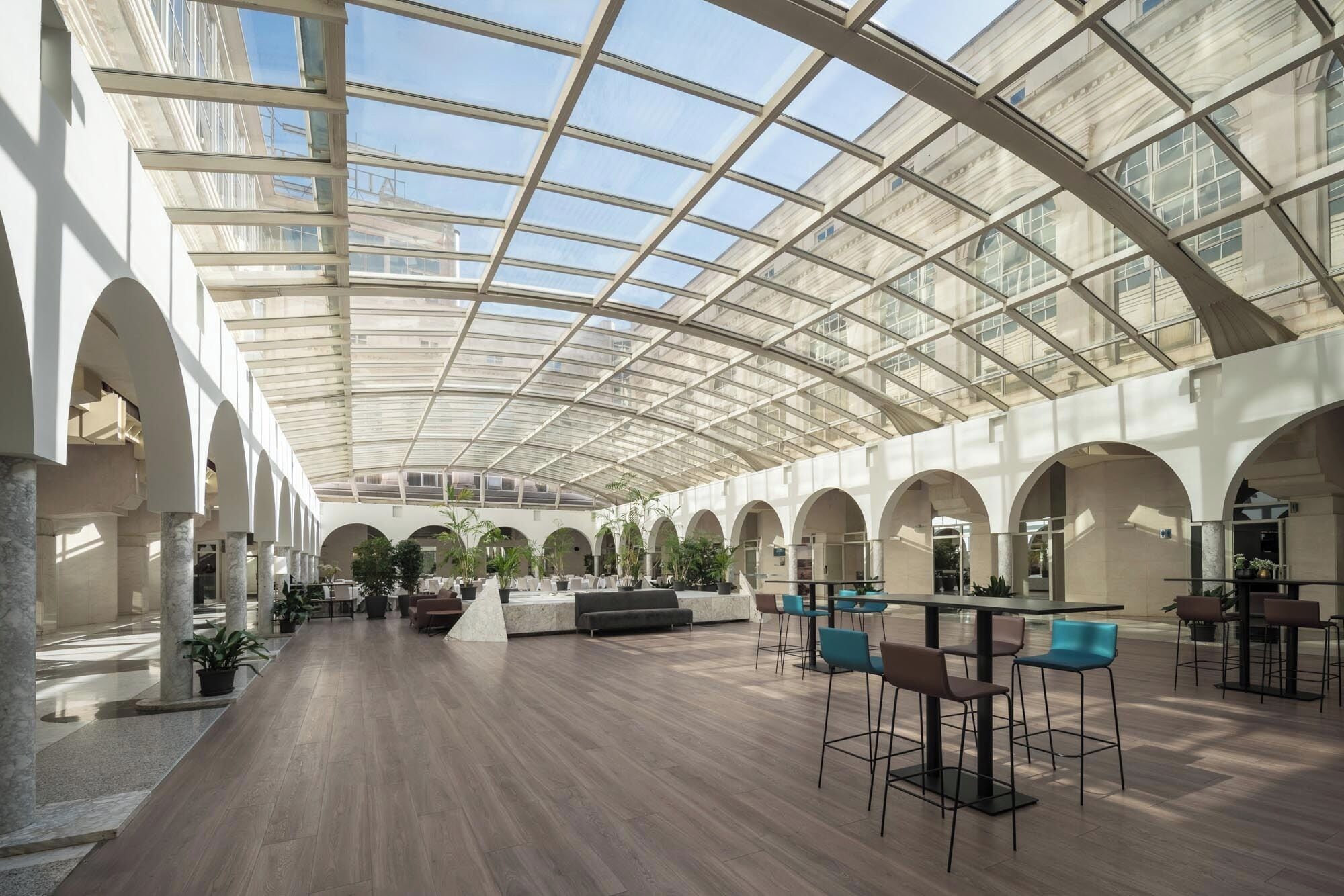 A spacious indoor area with a high ceiling and a glass roof, featuring a central seating area with chairs and tables, surrounded by columns and potted plants.