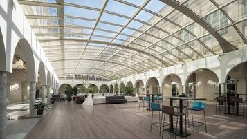 A spacious indoor area with a high ceiling and a glass roof, featuring a central seating area with chairs and tables, surrounded by columns and potted plants.