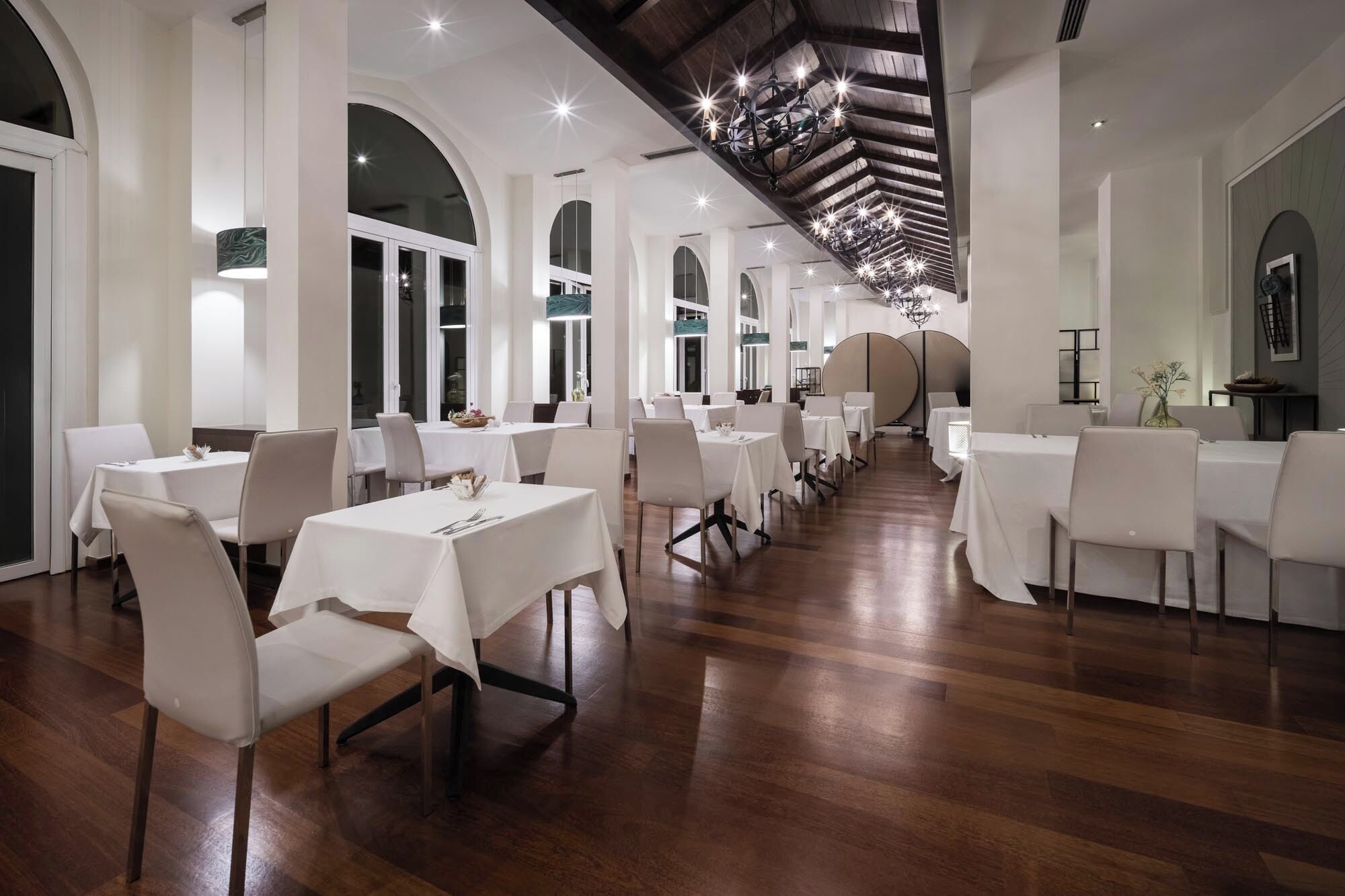 A spacious dining area with white tables and chairs, wooden flooring, and a high ceiling with exposed beams.