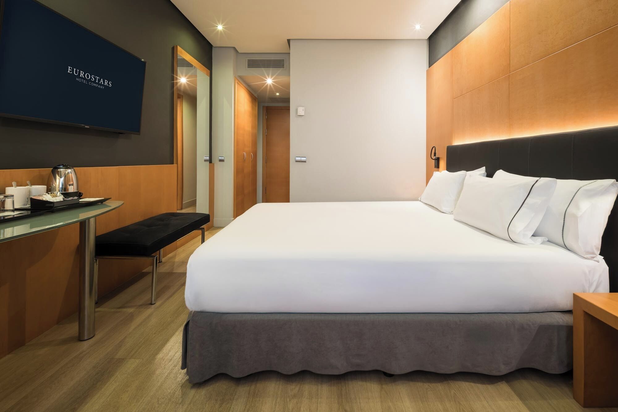 A hotel room with a bed, a desk with a coffee maker, and a TV mounted on the wall.