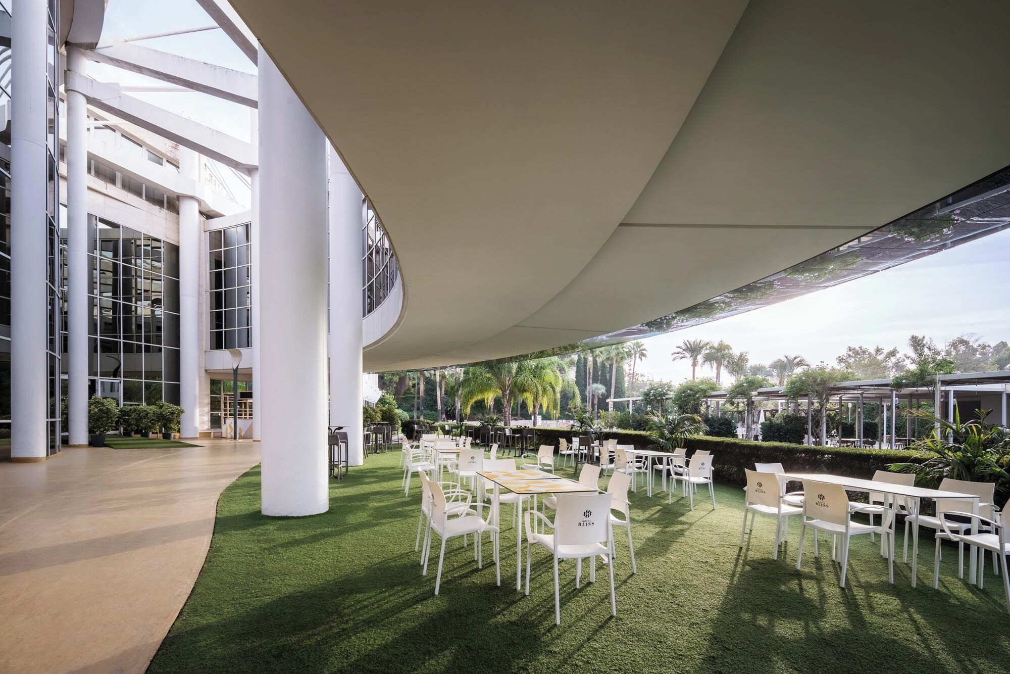 Un jardín interior moderno con sillas y mesas blancas, rodeado por un muro de cristal y un techo alto.