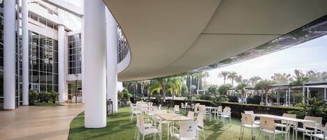 Un jardín interior moderno con sillas y mesas blancas, rodeado por un muro de cristal y un techo alto.