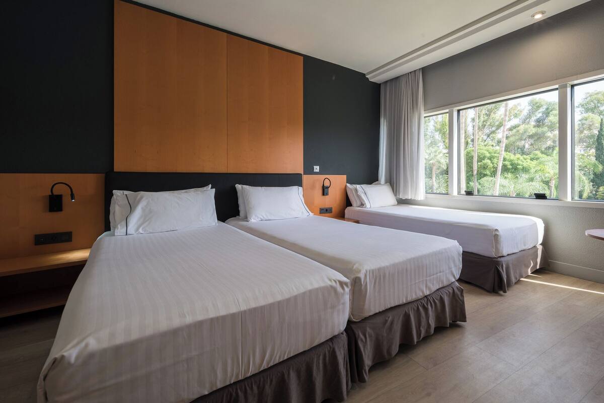 Habitación de hotel con dos camas, cabecera de madera, mesita de noche y ventana con vistas a zonas verdes.
