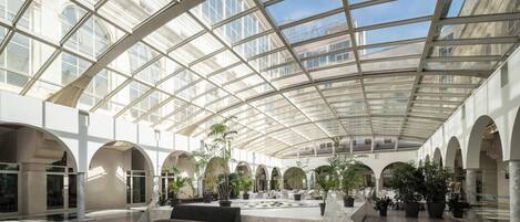 A spacious indoor area with a high ceiling and large windows, featuring a central seating area and multiple potted plants.