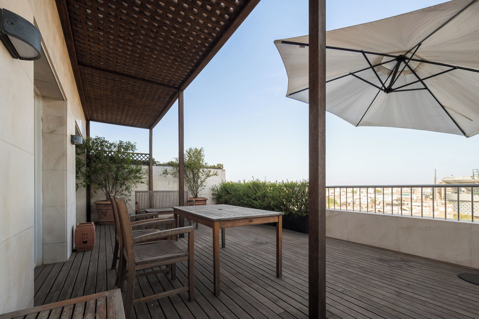 An outdoor patio with a wooden table and chairs, a large umbrella, and a view of the cityscape.