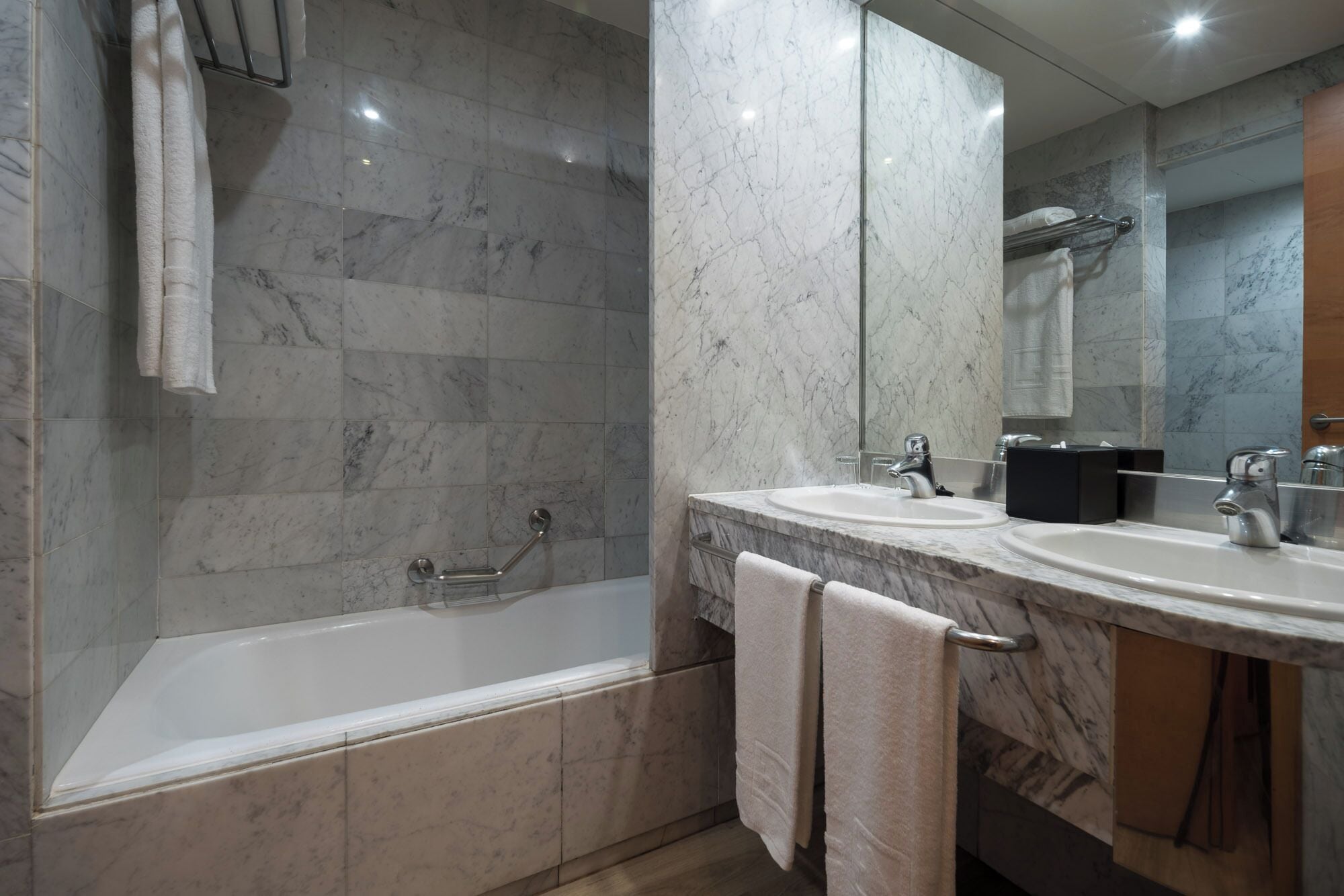A modern bathroom with a bathtub, two sinks, a mirror, and marble walls.