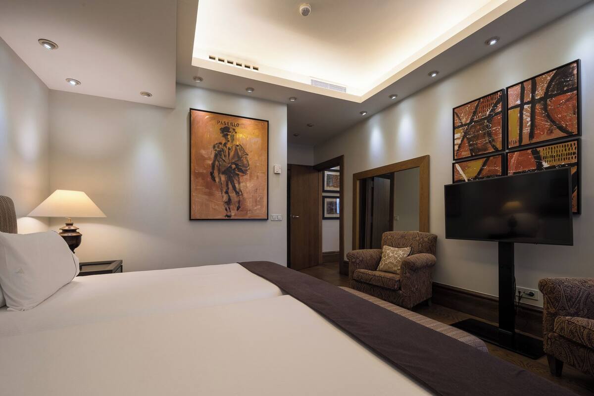 Habitación de hotel con una cama grande, dos sillones, un televisor y obras de arte en las paredes.