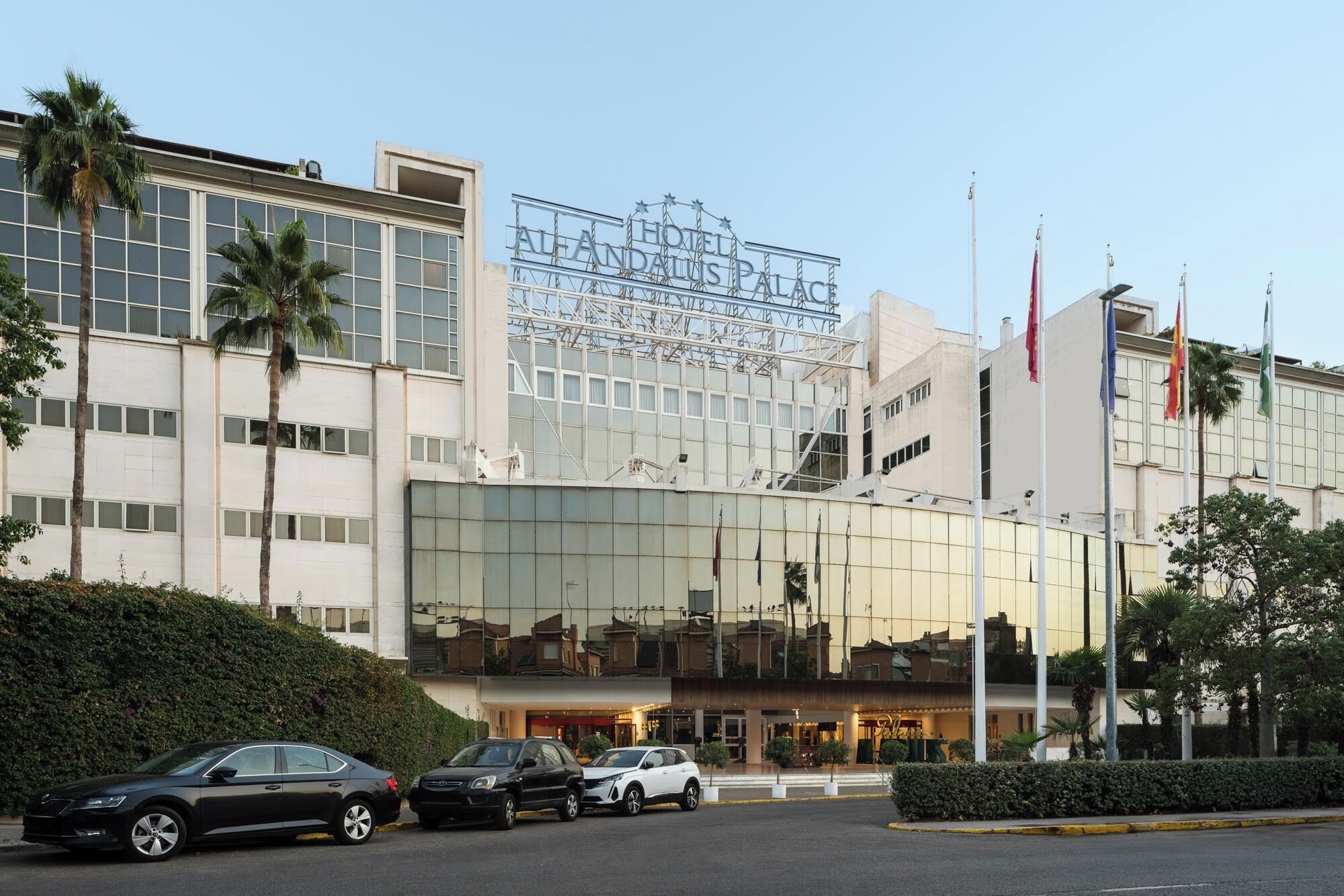 A modern hotel with a glass facade, palm trees, and flags.