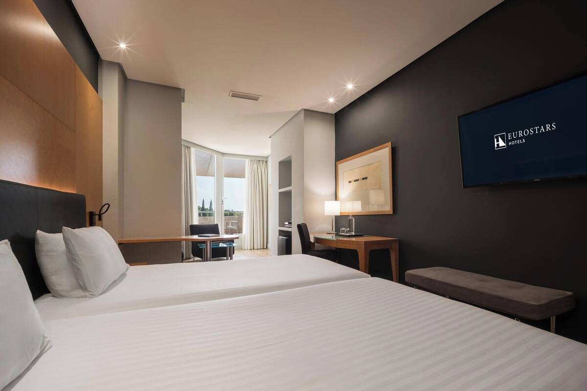 Una habitación de hotel con una cama grande, un escritorio, una silla, un televisor y un balcón.