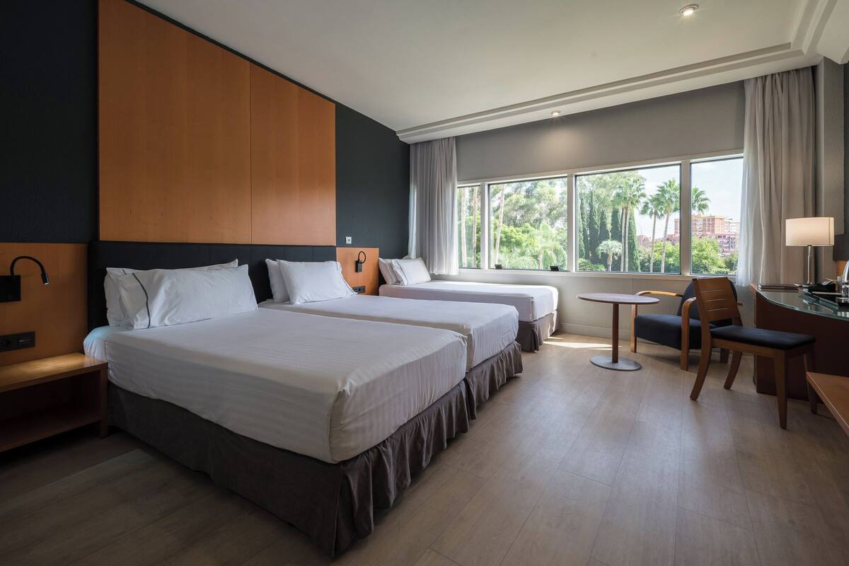 Habitación de hotel con dos camas, un escritorio con silla, una mesita y ventana con vistas a palmeras y edificios.