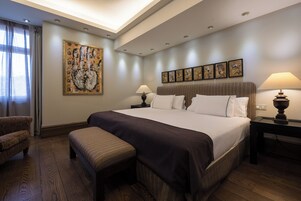 Une chambre d’hôtel avec un grand lit, deux tables de chevet avec des lampes, un banc, une chaise, une grande peinture abstraite et une série de petites œuvres d’art encadrées accrochées au mur.